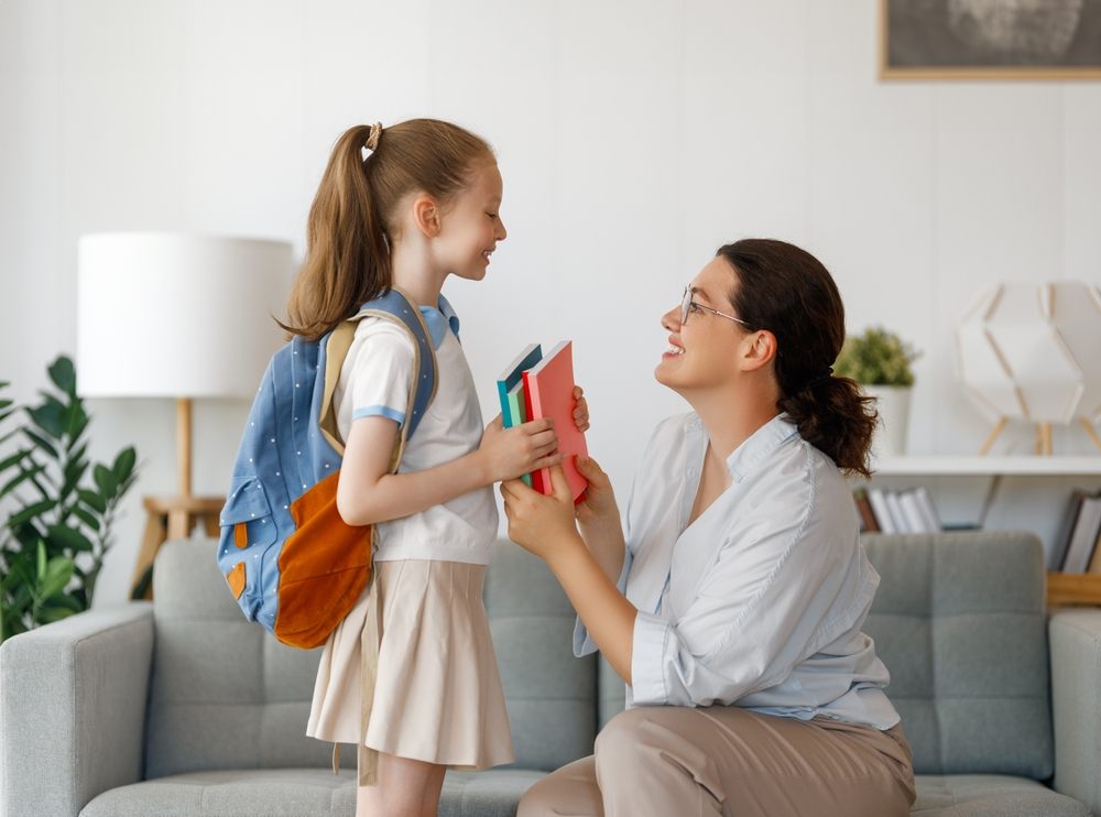Smiling mother gives school books to daughter with backpack before heading back to school