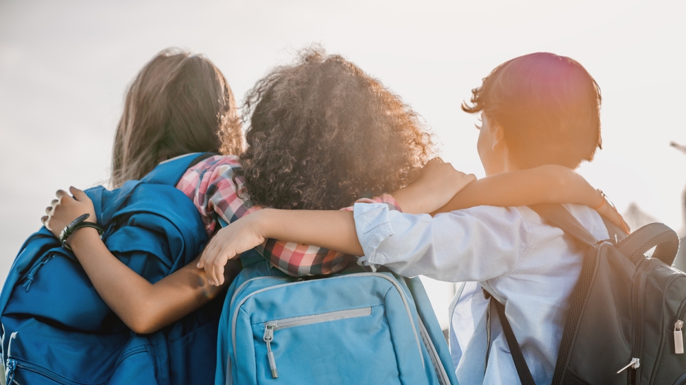Three children with backpacks embracing each other on first day of school, back-to-school season in Canada