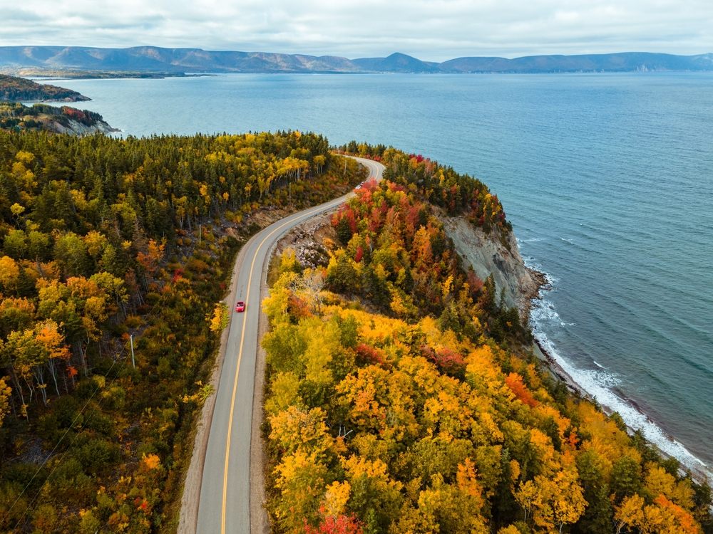Scenic coastal highway with red car driving through fall foliage in Canada, perfect for a summer road trip