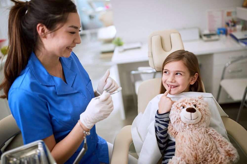 Smiling child patient with teddy bear at dental clinic receiving care from dental hygienist