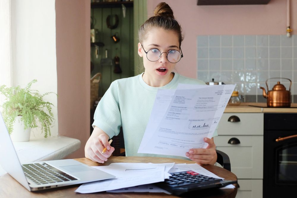 Surprised woman reviewing unexpected bills and expenses at home with laptop and calculator