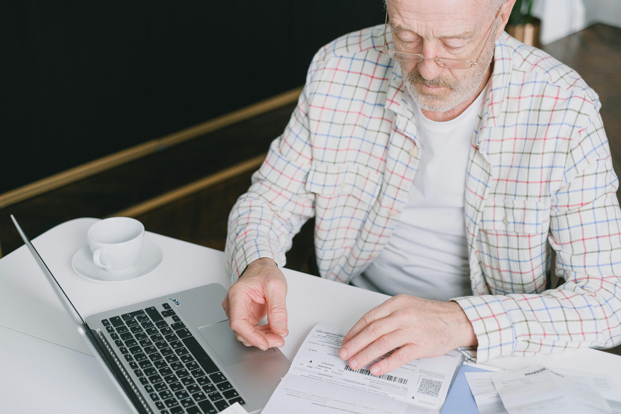 Elderly man with eyeglasses reviewing documents at a laptop. Indoor setting with natural light
