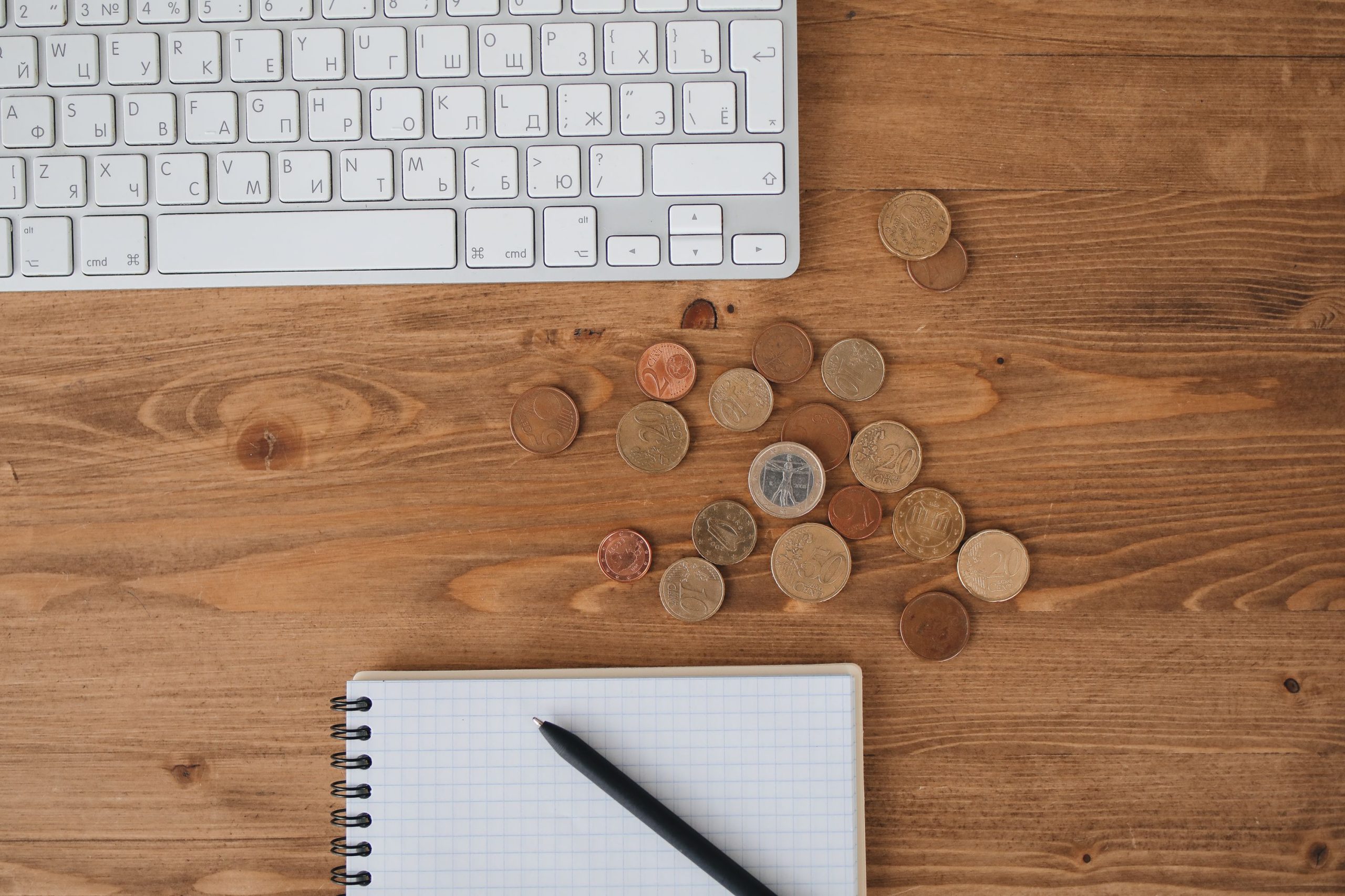 Scattered coins on a wooden desk alongside a keyboard and notebook, captured from above