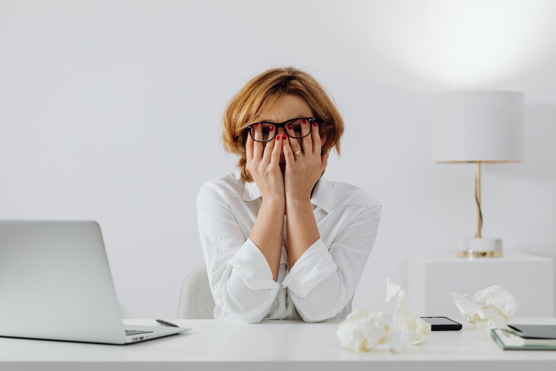 Woman with hands on face, sitting at desk with laptop and tissues, feeling stressed